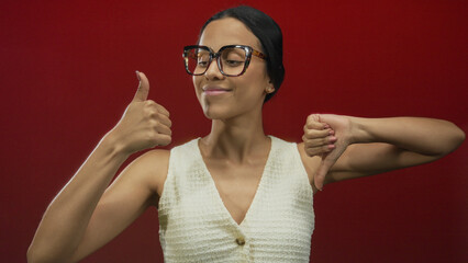 Hispanic woman wearing glasses showing thumbs up and down gestures against a vibrant red background.