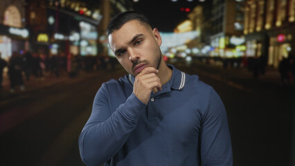 Young man wearing a blue polo shirt with hand on chin standing on busy city street at night;...