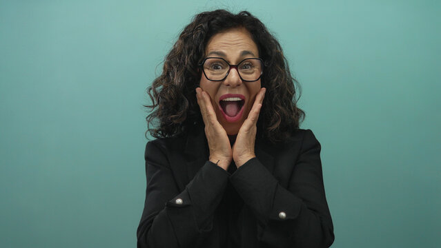 Woman expressing surprise with hands on cheeks against green wall background with shocked expression wearing black blazer and glasses middle aged hispanic female over isolated scene