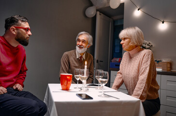 Cozy dinner setting featuring three adults enjoying conversation, highlighting familial togetherness and relaxation in warmly lit kitchen environment, complemented by beverages, candlelit atmosphere.