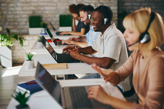Smiling African American male customer service agent works enthusiastically on his laptop and headset among diverse colleagues in a modern, busy call center office