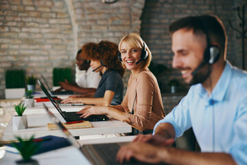 Smiling Caucasian female customer service agent looks directly at the camera while working on her laptop with diverse colleagues in a busy, modern call center office