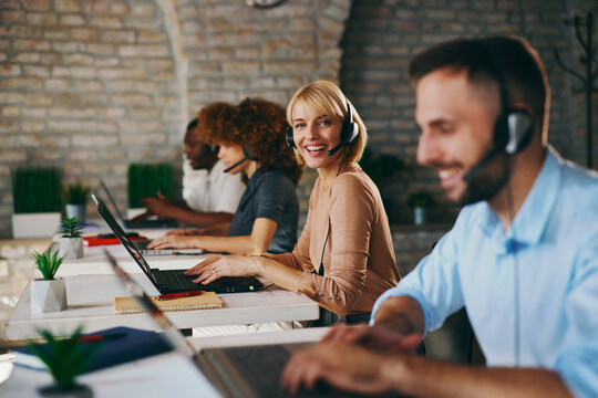 Smiling Caucasian female customer service agent looks directly at the camera while working on her laptop with diverse colleagues in a busy, modern call center office