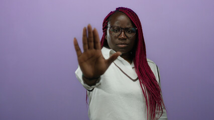 Woman with braids and glasses poses against purple wall making stop gesture wearing white shirt...
