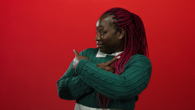Woman with pink braids in green sweater pointing sideways against red background looking thoughtful. - Powered by Adobe