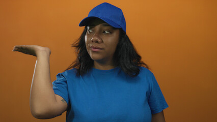 Woman wearing blue cap and shirt holds palm up with slight head tilt in bright orange studio; uncertain decision.
