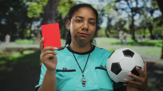 Referee in turquoise jersey shows red card and holds soccer ball in forest; authority decision fairness judgement.