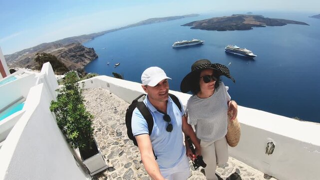 Romantic couple walking down a stone walkway on Santorini island sightseeing and caldera and city of Thera. The man is wearing a blue shirt and white shorts, and the woman is wearing a hat