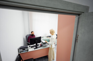 A bearded man sits at a computer while a blonde woman hands over files, showcasing collaboration in a clean, modern office with pastel walls and organized equipment.