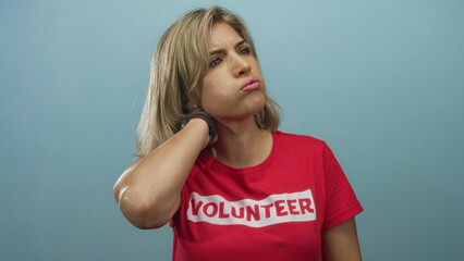 Woman volunteer holds her neck in studio against light blue wall with tense posture and visible blonde hair; discomfort.