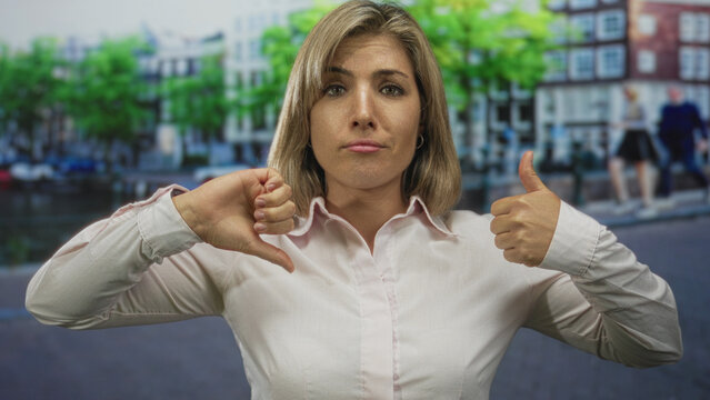 Blonde woman displays thumbs up and thumbs down on an amsterdam street by a canal; uncertainty conflict.