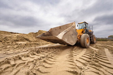 A large yellow bulldozer works diligently on a cloudy day, shaping the soil at a construction site surrounded by piles of dirt and tire marks on the ground