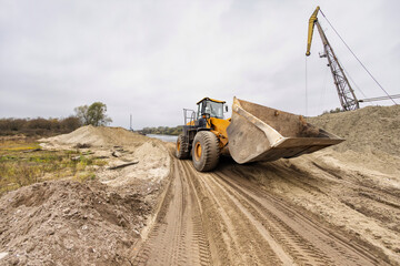 Fototapeta premium A giant loader works diligently, shifting soil along a riverbank, surrounded by mounds of dirt under a cloudy sky, signaling ongoing construction activity