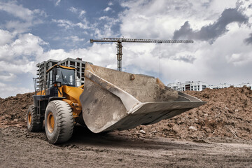 A powerful loader digs into the ground at a construction site, preparing for new buildings under a dramatic sky filled with clouds