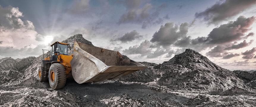 Large earth-moving equipment works tirelessly among towering piles of soil as the sun sets, casting shadows and illuminating the clouds overhead. Loader or bulldozer.