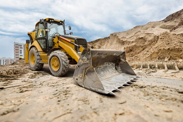 Fototapeta premium A yellow excavator moves through a construction site, skillfully digging and moving dirt against a backdrop of tall buildings and an overcast sky
