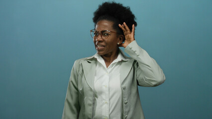 Fototapeta premium Woman listening intently with hand to ear against an isolated blue background, showcasing a thoughtful and curious expression in a business suit and glasses.