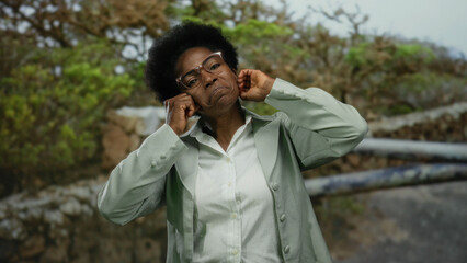 Woman wearing glasses plugs ears and makes a face in an outdoor park with trees, conveying frustration and a playful mood.