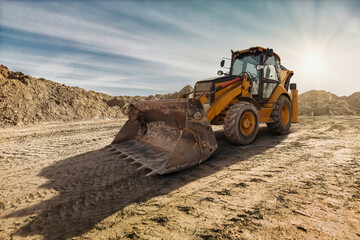 Obraz premium A yellow backhoe loader digs into the earth on a construction site as the sun sets, casting long shadows on the rugged ground