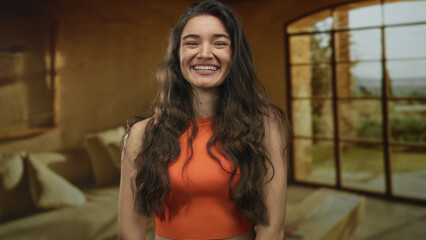 Fototapeta premium Woman wearing orange crop top with long wavy hair standing and pursing lips while laughing in sunlit living room; playful joy.