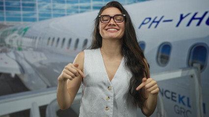 Young hispanic woman wearing glasses smiling and pointing finger at plane in airport terminal...