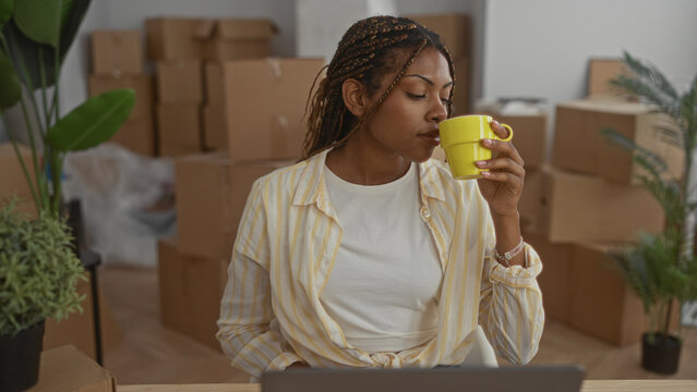 African american woman holding yellow cup and typing on laptop amid stacked moving boxes and houseplant in a house while settling in; focused.