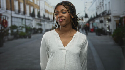 Fototapeta premium Woman smiling with braided hair and nose ring standing on a city street in front of shops wearing white blouse and relaxed posture; confidence.