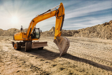 Heavy machinery digs into the dry soil of a construction site. The sun sets, casting warm hues over...