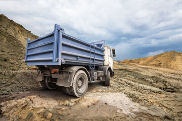 A large dump truck drives along uneven ground, leaving tire tracks in the dirt, as dark clouds...