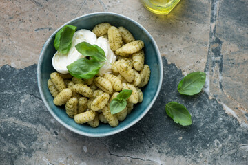 Bowl of potato gnocchi with basil oil, torn mozzarella and fresh basil, horizontal shot on a grey and beige granite background, flat lay