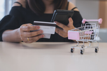 Woman making online payment with credit card and smartphone, mini shopping cart on table symbolizing e-commerce, digital shopping, secure transaction and modern retail lifestyle.