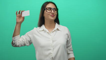 Naklejka premium Woman holding white card and pointing finger in studio teal backdrop, wearing glasses and white shirt, smiling toward camera; confidence.