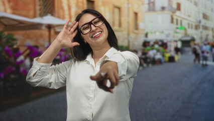 Woman pointing finger to camera on street terrace wearing glasses and smiling in white blouse; joy playful energy.