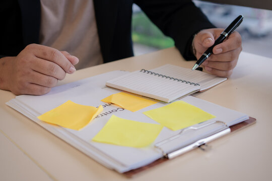 Close-up of a businessperson writing notes in a lined notebook with sticky notes and documents on a clipboard, planning tasks and organizing work for productivity and focus.