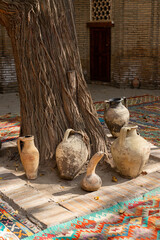 Ancient tree trunk surrounded by clay jugs and colorful woven carpets in a historical courtyard in...