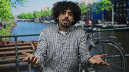 Man gesturing open hands with bare hands near bicycle on a street by a canal in amsterdam wearing...