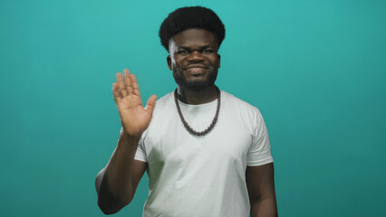 Young man smiling and waving hand, wearing white t shirt and beaded necklace in studio with teal...