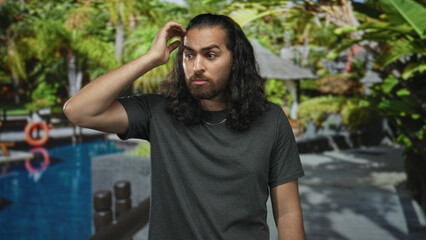 Man scratching head with hand beside pool railing at resort building walkway with palm trees and lifebuoy visible; puzzlement indecision vacation.