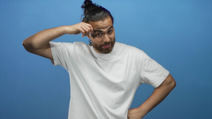 Naklejka premium Man with hand to forehead and bare forearm in blue studio, wearing a white t shirt and bun hairstyle while grimacing slightly; embarrassment fatigue.