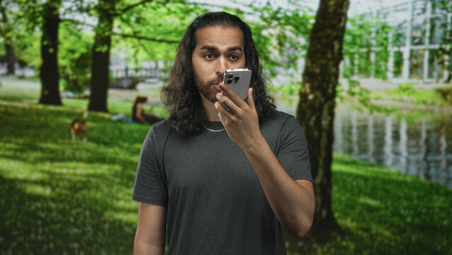 Young man with long hair and beard speaking into smartphone held in right hand in forest by a riverside pond; surprise voice assistant.