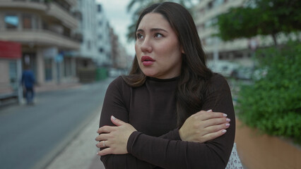 Fototapeta premium Hispanic woman holds temples on city street lined with apartments and parked cars while wearing long dark top; discomfort.
