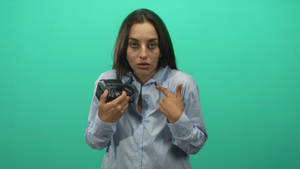 Woman holding camera and pointing finger to chest in green studio wearing light blue shirt and holding lens cap; confusion uncertainty.