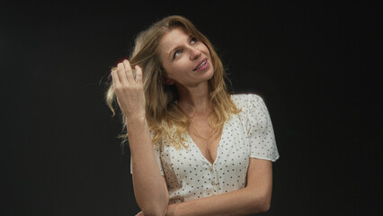 Fototapeta premium Woman caucasian in polka dot blouse touches hair revealing bare neckline in black studio while gazing upward with soft smile; thoughtful calm.