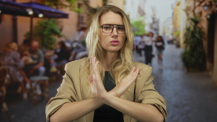 Obraz premium Blonde woman on terrace crossing arms in an x sign, rejecting idea in an outdoor city café setting.