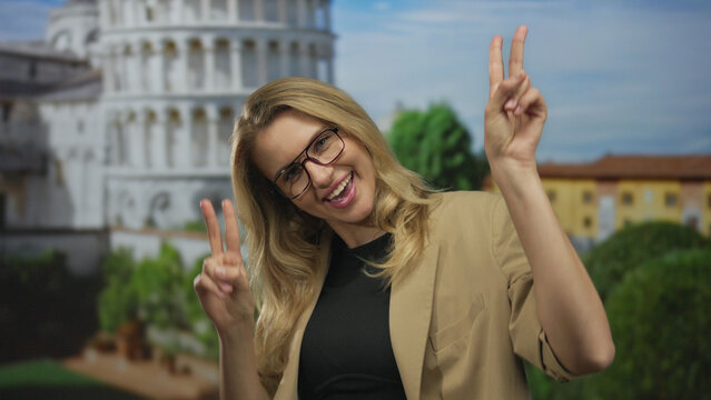 Woman smiling outdoors in pisa with blonde hair and glasses, making a peace sign near the iconic leaning tower on a sunny day.