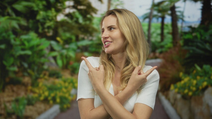 Young blonde woman pointing playfully in a lush green outdoor park setting with a cheerful expression amidst vibrant foliage and sunshine.