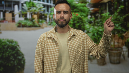 Obraz premium Man pointing finger upward on a city street with potted plants and concrete planters, wearing a beige checked shirt and neutral sweater; confidence.
