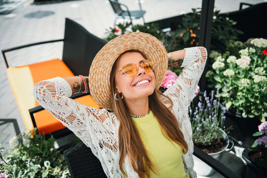 Relaxed stylish woman wearing sun hat smiles in bright outdoor cafe garden enjoying summer fashion and leisure on a sunny day - Powered by Adobe
