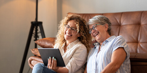 Portrait of happy mature blonde curly woman sitting on the floor close to her elderly mother having fun while looking together at digital tablet. The scene captures affection and companionship