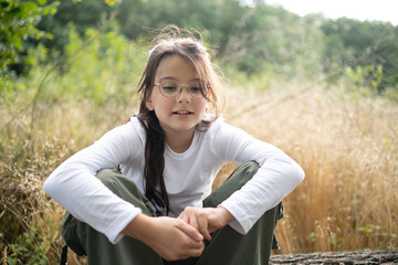 A smiling girl in glasses sitting in the forest, looking down thoughtfully with a gentle expression. Blurred golden grass and sky in the background, soft natural light, copy space. 